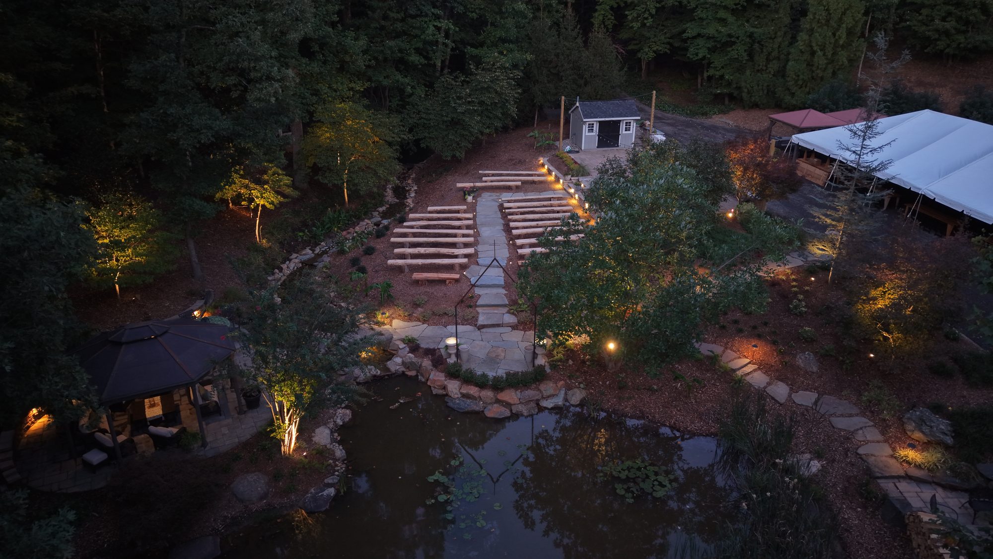 Aerial view at dusk of a farm wedding venue with a lit stone path, wooden ceremony benches, a pond, and a white reception tent