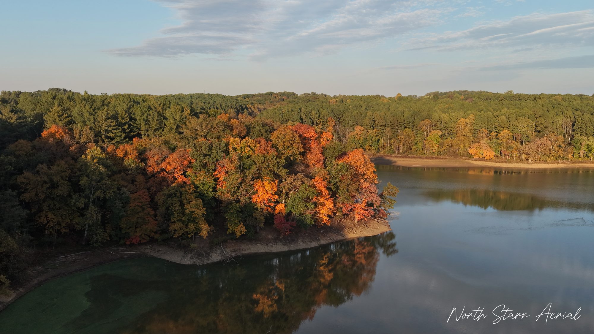 Aerial view of a peninsula of orange and red autumn trees reflected in the calm water of Liberty Reservoir at golden hour